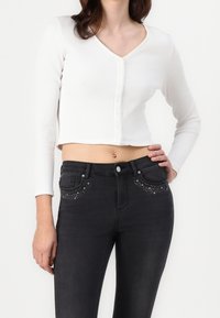 Gerippte weiße, kurz geschnittene Strickjacke mit langen Ärmeln und Knopfverschluss, kombiniert mit hochsitztender schwarzer Jeans mit Nietenakzenten an den Taschen.