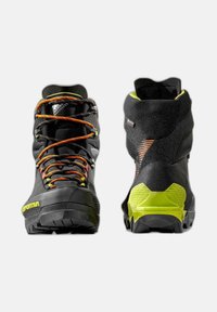 Zwarte wandelschoenen met een bovenwerk van gemengde materialen, feloranje en gele veters, een gestructureerde zwarte hak en een robuuste zwarte en gele zool.