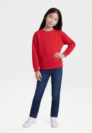 Jauna meitene ar gariem tumšiem matiem, tērpusies sarkanā sporta džemperī, zilās džinsu biksēs un baltos sporta apavos, stāv ar vienu roku uz gurna.