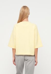 Īsām piedurknēm dzeltena džemperis no mīksta auduma ar vaļēju piegriezumu un taisnu apmali, valkāts kopā ar gaiši pelēkām džinsa biksēm.