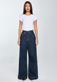 Široke temno modre jeans hlače z kontrastnim šivom, visokim pasom in gumbom, v kombinaciji z oprijeto belo majico.