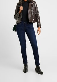 Brun faux läder bikerjacka, svart topp, mörkblå skinny jeans och svarta ankle boots med lätt klack. Grön axelväska synlig som detalj.