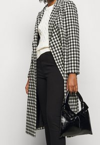 Abrigo largo a cuadros en blanco y negro, suéter de punto trenzado blanco, pantalones negros, sosteniendo un bolso brillante negro con un detalle de lazo.