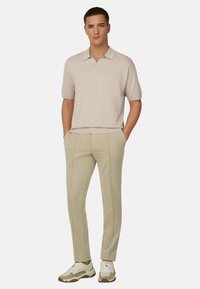 Jeune homme debout, les mains dans les poches, portant un polo beige, un pantalon beige avec des coutures avant, et des baskets blanches, sur un fond blanc.