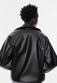 Črna jakna iz umetne usnjene tkanine z teksturirano površino, oversize krojem, širokim ovratnikom in spuštenimi rameni. Zadnji pogled prikazuje drapirano zasnovo oblačila.