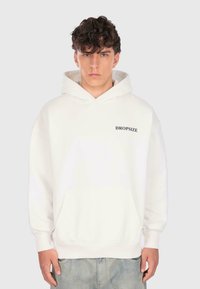 Junger Mann mit dunklem, lockigem Haar trägt einen weißen Hoodie mit dem Aufdruck "DROPSIZE" auf der Brust und hellblauen Jeans, steht vor einem schlichten Hintergrund.