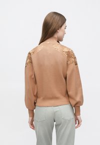 Bruine pullover trui met kanten details op de schouders, geribbelde manchetten en zoom, en een afgeronde halslijn. Draagt over lichtgrijze spijkerbroek.