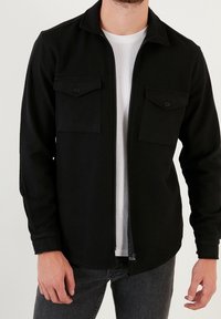 Sort lynlåsjakke med krave, to brystlommer og knaplukning. Fremstillet af et tekstureret stof, båret over en hvid skjorte.