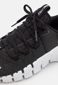 Scarpa sportiva nera con tomaia in mesh, lacci elastici e suola in gomma bianca con motivo testurizzato. Dettagli in nero e grigio.