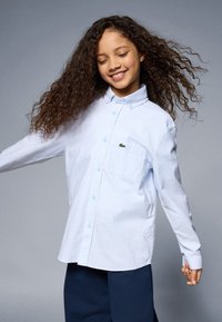 Camicia bianca a maniche lunghe con design a bottoni, dotata di una tasca sul petto e un piccolo logo verde. Il tessuto ha una consistenza liscia.