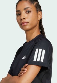 Tricou sportiv negru cu mâneci scurte, având trei dungi albe pe mâna stângă și un mic logo pe față. Textură moale.