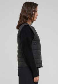Gilet matelassé noir avec un col montant, des boutons-pression sur les côtés et une texture douce, porté sur un pull noir à manches longues. Vue de côté.