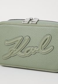 Borsa in pelle verde con texture, dotata di un logo "Karl" pronunciato in finitura rialzata a contrasto e dettagli in metallo argentato. Chiusura a doppia cerniera.