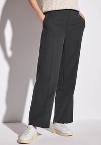 Pantaloni larghi grigio scuro con una texture liscia, dotati di tasche laterali, abbinati a sneaker bianche con suola crema.