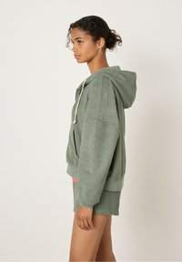 Felpa verde in spugna con cappuccio a coulisse, vestibilità ampia e orlo a coste, abbinata a pantaloni corti coordinati. Tessuto testurizzato con un aspetto morbido.