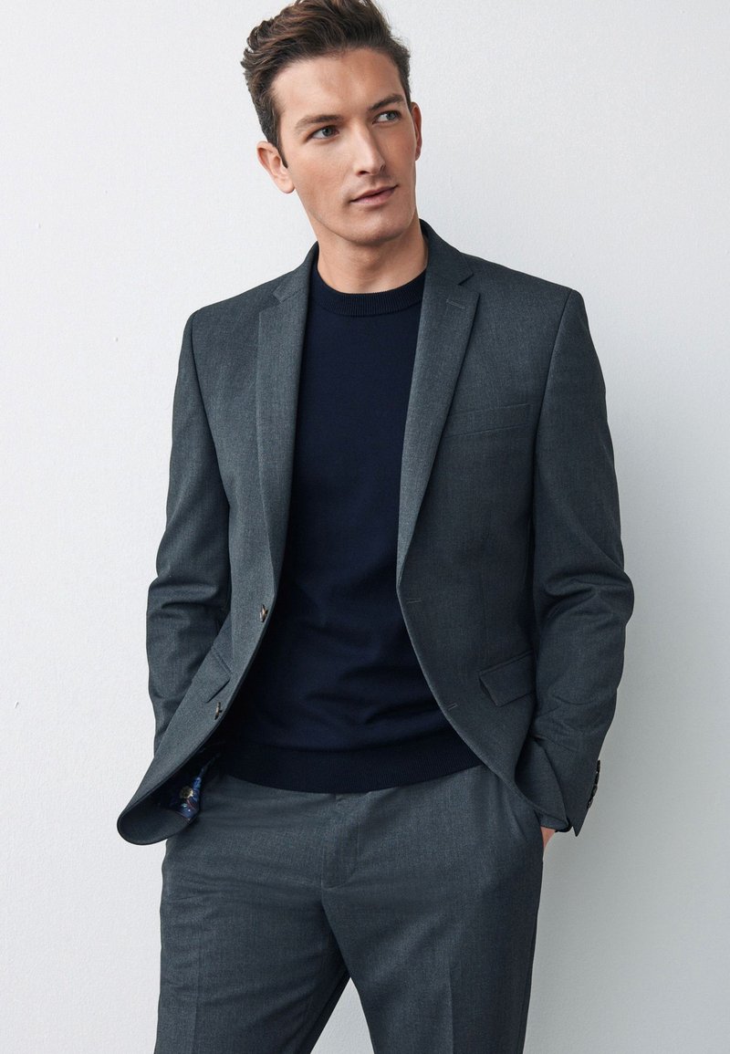 Grå jakkesæt med strukturerede revers, enkeltradet design og subtilt ternet mønster. Båret over en navy sweater med struktureret stof.