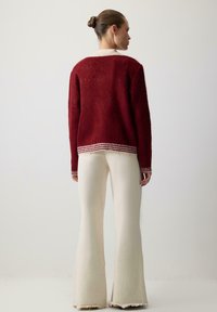 Czerwona sweter dziany z długimi rękawami i paskowanym dołem, w połączeniu z off-white spodniami w dzwon, wszystko zaprezentowane na neutralnym tle.