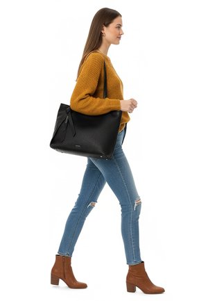 Frau geht und trägt einen senffarbenen Pullover, zerrissene Skinny-Jeans, braune Stiefeletten und trägt eine große schwarze Umhängetasche.