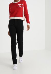 Osoba idąca w czerwonym swetrze z białym wykończeniem i literami "TJ", czarnych dżinsach i białych sneakersach na jednolitym białym tle.