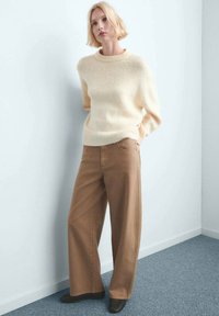 Pull en tricot couleur crème avec un col rond, associé à un pantalon large couleur marron clair et des chaussures marron foncé, sur un fond bleu pâle.