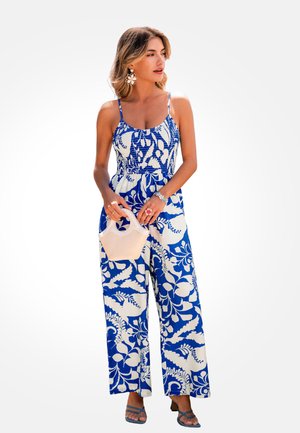 Vrouw in een blauw-witte jumpsuit met bloemenprint, met een kleine beige handtas, grote bloemoorbellen en sandalen, staand tegen een effen achtergrond.