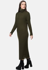 Olivgrünes, geripptes, gestricktes Rollkragenkleid mit langen Ärmeln, Seitenschlitz und schwarzen Plateaustiefeln bis zum Knöchel. Glatte Textur und figurbetontes Design.