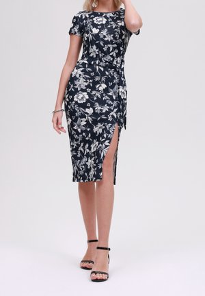 Kobieta w czarno-białej sukience o długości do kolan z kwiatowym wzorem, z bocznym rozcięciem i krótkimi rękawami, w połączeniu z czarnymi sandałami na kostkę na obcasie.