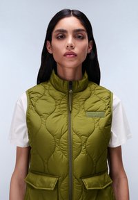 Gequilte olijfgroene vest met hoge kraag, rits aan de voorkant en twee voorzakken. Heeft subtiele branding en een veelzijdig, gewatteerd ontwerp.