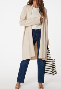 Beżowy kardigan zapinany na wierzchu, noszony na jasnej bluzce, w połączeniu z niebieskimi dżinsami i przezroczystymi sandałami na słupku. Czarna torba w białe paski.