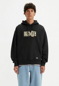 Black hoodie made of soft fabric featuring a large, gold, block-letter logo on the front. Includes a kangaroo pocket and drawstring hood.