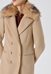 Cappotto beige doppiopetto con finitura testurizzata, dotato di un colletto in pelliccia morbida nei toni del marrone e due tasche laterali.