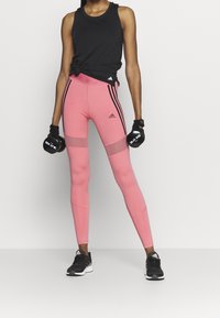 Różowe legginsy sportowe z czarnymi paskami po bokach, w połączeniu z czarną bezrękawnikową bluzką. Czarne rękawiczki i buty sportowe dopełniają stylizację.