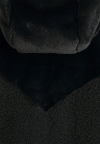 Primo piano di un tessuto scuro e strutturato con superfici morbide e pelose di varie consistenze e dettagli di cucitura che formano un motivo sovrapposto.