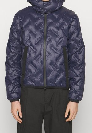 Personne portant une doudoune matelassée bleu marine avec une fermeture éclair noire et des panneaux latéraux, associée à un pantalon noir ample, debout devant un fond uni.