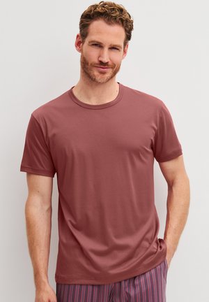 Man met krullend haar en baard, die een effen mauve t-shirt en gestreepte broek draagt, staand tegen een effen lichte achtergrond.