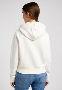 Weißer Kapuzenpullover mit lockerer Passform, ausgestattet mit einer Kordelzugkapuze und gerippten Bündchen. Kombiniert mit blauen Jeans, minimalistischem Design und weicher Textur.