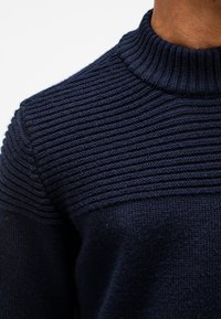 Marineblauwe gebreide trui met een hoge ribkraag en gestructureerde horizontale stiksels op de schouders. Zachte, middelzware stof.