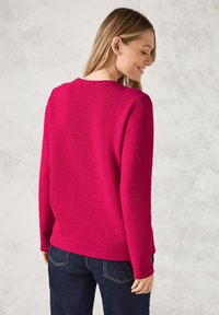 Magenta, texturerad tröja med fiskbensmönster, långa ärmar och rund halsringning, tillsammans med mörka denimjeans.