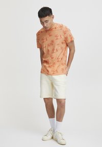 En oransje t-skjorte med korte ermer og et subtilt mønster, kombinert med lysegule shorts. Fottøyet har en kremfarge med teksturerte detaljer.