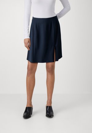 Vrouw draagt een zwarte rok tot op de knie met een zijsplit, een wit topje met lange mouwen en zwarte slip-on schoenen met hak, staand voor een effen achtergrond.