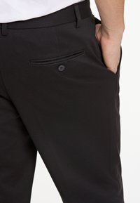 Schwarze maßgeschneiderte Hose mit einer Seitentasche und Knopfverschluss sowie einer glatten, strukturierten Oberfläche. Der moderne, figurbetonte Schnitt betont die Silhouette.