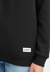 Felpa nera con orlo a costine, realizzata in tessuto liscio. Presenta un'etichetta bianca con il logo 'oxmo' vicino al fondo.