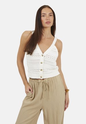 Vrouw draagt wit gebreid topje zonder mouwen met knopen en beige losse broek met trekkoord, poseert met hand in zak tegen een effen achtergrond.