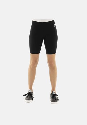 Benen met zwarte knielange sportshorts en zwarte sneakers met witte zolen op een effen lichte achtergrond.