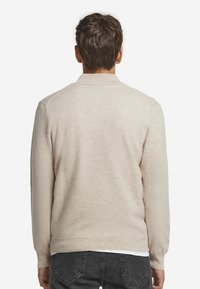 Jasno beżowy sweter, dziany teksturą, z długimi rękawami i ściągaczem na dole. Brak widocznych wzorów ani ozdób. Widok z tyłu z gładkim wzorem.