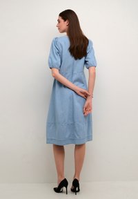 Gaiša zila denim midi kleita ar pūkainām piedurknēm, savilkuma detaļām pie aprocēm un pakāpenisku apakšmalu. Valkāta ar melniem augstiem zābakiem.