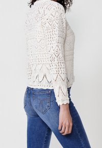 Bílý pletený svetr se složitými vzory s otevřenou prací, dlouhými rukávy a vroubkovaným lemem, v kombinaci s přiléhavými modrými džínami s kapsami na zadní straně.