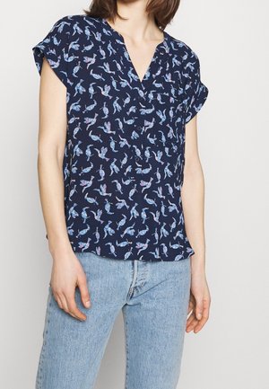 Kobieta ubrana w granatową bluzkę z krótkim rękawem z nadrukiem ptaków, zestawioną z jasnoniebieskimi wysokim stanem jeansami.