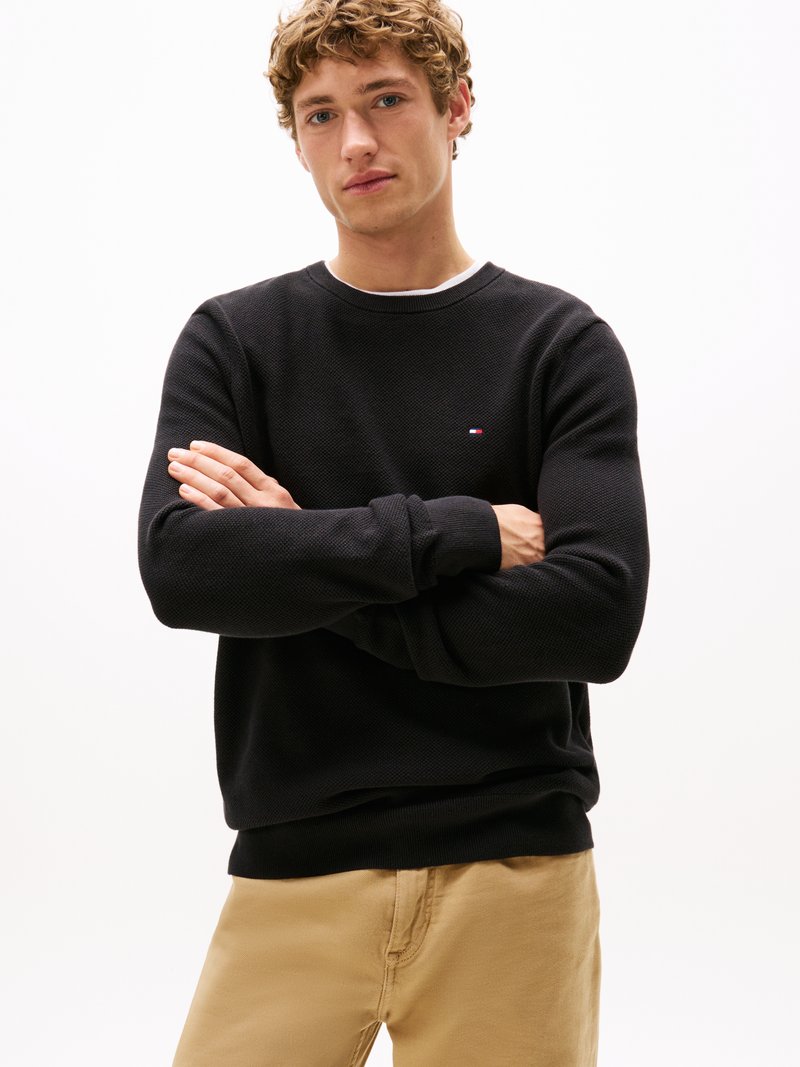 Sweat-shirt noir à col rond avec des poignets et un ourlet côtelés, affichant un petit logo sur la poitrine. Tissu texturé associé à un pantalon beige.