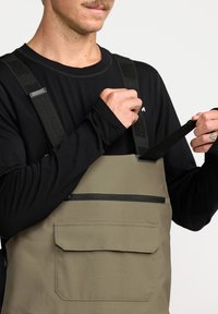 Kaki waterdichte bretels met verstelbare zwarte banden, voorzien van een ritszak aan de voorkant en een flapzak, ontworpen voor duurzaamheid en functionaliteit.